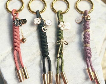 May include: Four keychains with gold-colored rings and accents. Each keychain features a different color of braided cord: pink, black, green, and purple. Charms include flowers, smiley faces, hearts, and initial letters. The keychains have gold-colored cylindrical end caps.