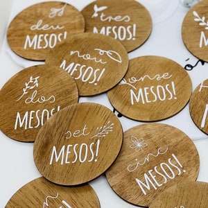 Puede incluir: Un conjunto de diez círculos de madera con texto blanco y diseños florales. Cada círculo presenta un número del uno al diez en español, seguido de la palabra "MESOS".