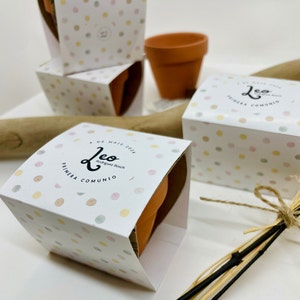 Puede incluir: Una caja blanca con un patrón de lunares de colores y el texto "Leo Primera Comunio" y "4 de Maig 2024" impreso en ella. La caja contiene una pequeña maceta de terracota.