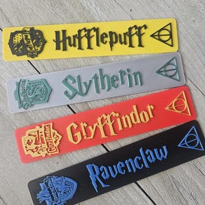 May include: Four Harry Potter house bookmarks with the house crest and name. The bookmarks are yellow, gray, red, and black. The house names are Hufflepuff, Slytherin, Gryffindor, and Ravenclaw.