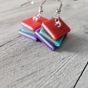 May include: A pair of book-shaped earrings with a stack of four books in red, purple, blue, and white. The earrings are made of polymer clay and have silver hooks.