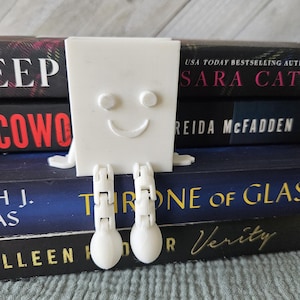 Pode incluir: Um separador de livros branco impresso em 3D com a forma de um rosto sorridente com braços e pernas. O separador de livros está segurando uma pilha de livros. O livro de cima é intitulado "Sara Cat" de Reida McFadden. O segundo livro é intitulado "Throne of Glass" de A.J. Verity. O terceiro livro é intitulado "Fallen" de Lauren