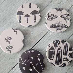 May include: Set of six round car coasters with different designs. The coasters feature black and white illustrations of mushrooms, butterflies, snakes, crystals, and constellations on a beige background.