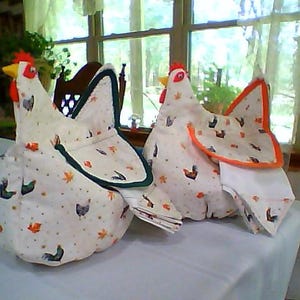 May include: Two white fabric chicken-shaped tissue holders with orange and green accents. The holders are sitting on a white tablecloth.