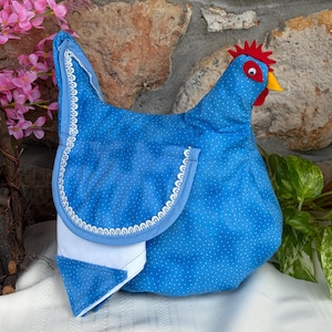 May include: A blue fabric chicken-shaped item with a white polka dot pattern. It features a red felt comb, a yellow beak, and a googly eye. The wing is accented with white lace trim, and a white fabric section is visible at the bottom edge.