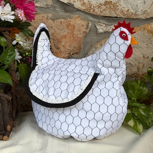 May include: A fabric chicken-shaped tea cozy featuring a white background with a black hexagonal wire fence pattern. It has a red felt comb, a yellow beak, a red wattle, and a googly eye. The wing is accented with white lace trim.