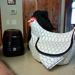 May include: A white and black chicken-shaped toaster cover with a red comb and yellow beak. The cover has a honeycomb pattern and black trim. A black and red toaster is to the left of the cover.