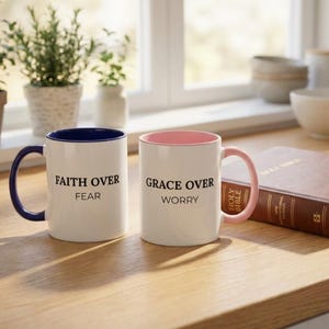 May include: Two white ceramic mugs with inspirational quotes. One mug has a navy blue handle and rim with the text "FAITH OVER FEAR." The other mug has a pink handle and rim with the text "GRACE OVER WORRY."