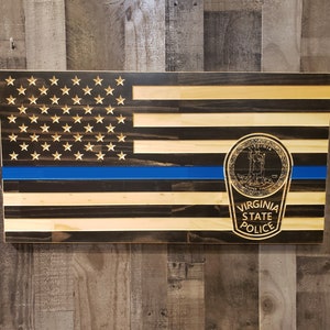 May include: A wooden American flag with a blue line and the Virginia State Police emblem. The flag is made of dark wood with light wood stripes. The emblem is gold and features the state seal of Virginia.