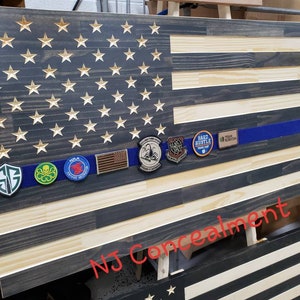 Op de afbeelding: Een Amerikaanse vlag van hout met een blauwe lijn in het midden. De vlag is versierd met verschillende patches, waaronder een "Hard Hustle"-patch, een "Team Hudson"-patch en een "SG"-patch. De vlag wordt getoond op een houten ezel en heeft de tekst "NJ Concealment" in rood onderaan.