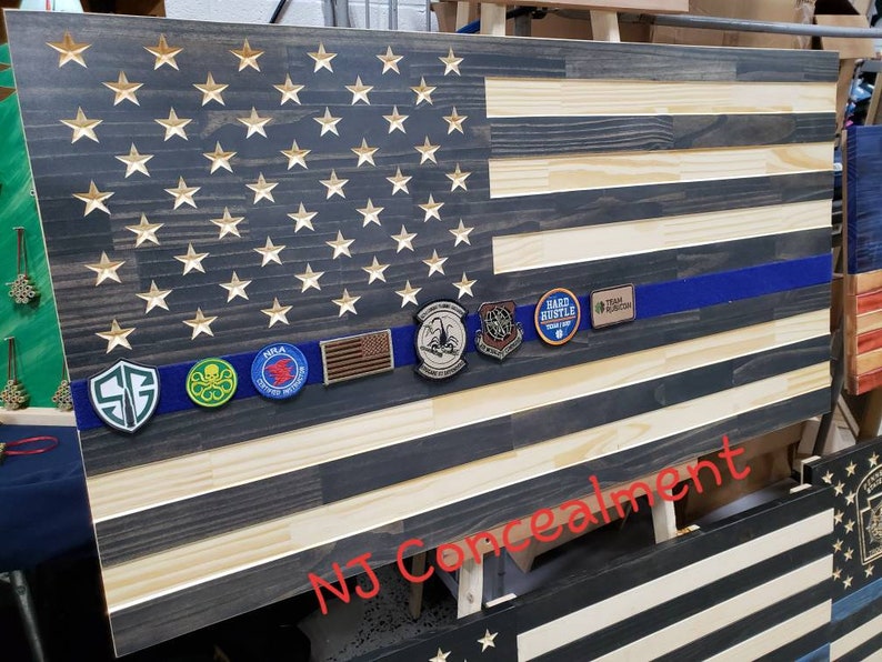 Op de afbeelding: Een Amerikaanse vlag van hout met een blauwe lijn in het midden. De vlag is versierd met verschillende patches, waaronder een "Hard Hustle"-patch, een "Team Hudson"-patch en een "SG"-patch. De vlag wordt getoond op een houten ezel en heeft de tekst "NJ Concealment" in rood onderaan.