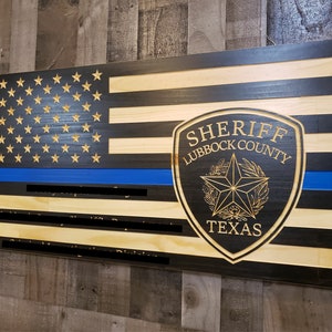 May include: Wooden American flag with a blue line, a black sheriff's badge with the text "Sheriff Lubbock County Texas" in gold lettering, and a gold star.