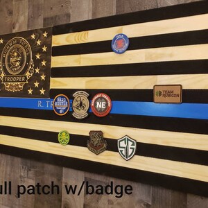 Op de afbeelding: Een Amerikaanse houten vlag met een blauwe lijn, met verschillende patches en een badge van de Florida Highway Patrol Trooper. De patches omvatten "Hard Hustle", "Northeast Territory", "Team Rubicon" en "S.G.".