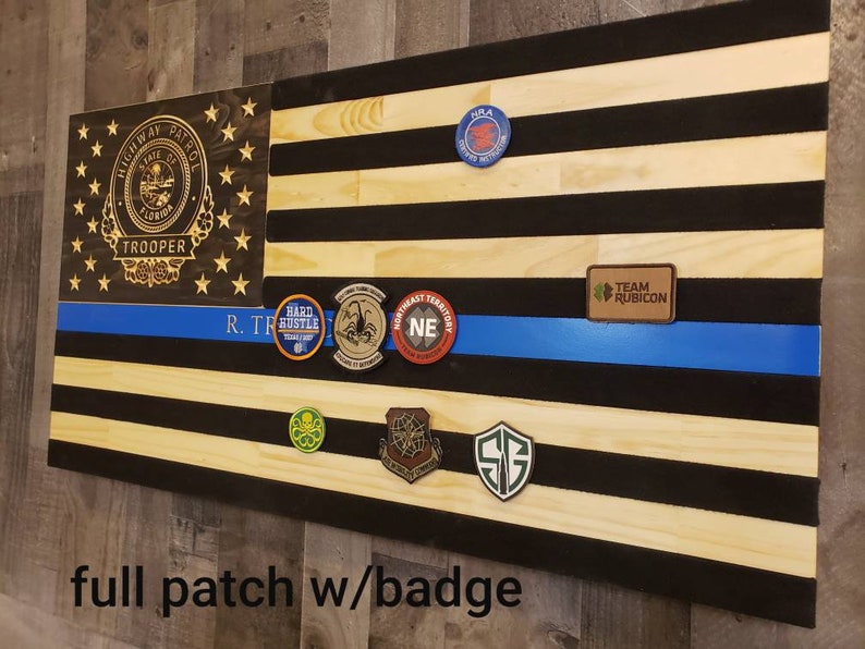 Op de afbeelding: Een Amerikaanse houten vlag met een blauwe lijn, met verschillende patches en een badge van de Florida Highway Patrol Trooper. De patches omvatten "Hard Hustle", "Northeast Territory", "Team Rubicon" en "S.G.".