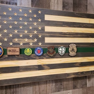 Op de afbeelding: Een Amerikaanse vlag van hout met een donker gebeitst oppervlak en lichte houten strepen. De vlag heeft een groen lint met verschillende patches, waaronder "Hard Hustle", "Team Rubicon", "NILA", "SF", en een schorpioenpatch.