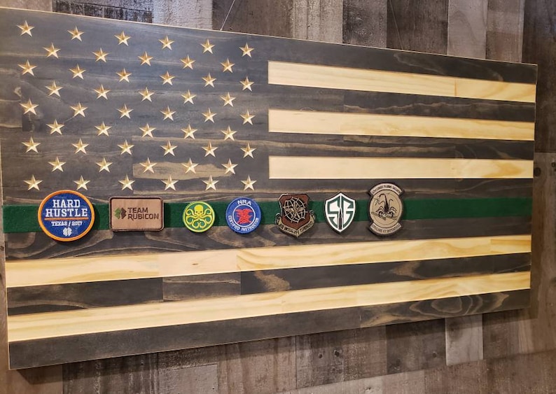 Op de afbeelding: Een Amerikaanse vlag van hout met een donker gebeitst oppervlak en lichte houten strepen. De vlag heeft een groen lint met verschillende patches, waaronder "Hard Hustle", "Team Rubicon", "NILA", "SF", en een schorpioenpatch.