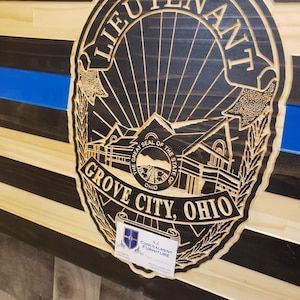 K&ouml;nnte beinhalten: Amerikanische Holzflagge mit blauem Streifen und geschnitztem Polizeiausweis mit der Aufschrift "LIEUTENANT GROVE CITY, OHIO". Die Flagge hat schwarze und nat&uuml;rliche Holzstreifen. Eine kleine Visitenkarte befindet sich in der unteren rechten Ecke.