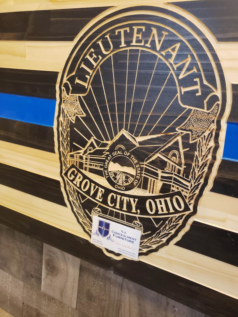 K&ouml;nnte beinhalten: Amerikanische Holzflagge mit blauem Streifen und geschnitztem Polizeiausweis mit der Aufschrift "LIEUTENANT GROVE CITY, OHIO". Die Flagge hat schwarze und nat&uuml;rliche Holzstreifen. Eine kleine Visitenkarte befindet sich in der unteren rechten Ecke.