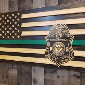 May include: A wooden American flag with a green stripe and a CBP Border Patrol badge. The badge is engraved with the words "CBP Border Patrol", "Department of Homeland Security", "Supervisory Patrol Agent", and "U.S."