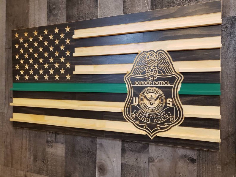 May include: Wooden American flag with a green line and a CBP Border Patrol badge. The flag features laser-cut stars and alternating black and natural wood stripes. The badge is detailed with text and an eagle.