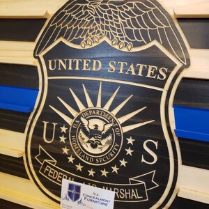 May include: Wooden wall art featuring a carved United States Federal Air Marshal badge. The badge is black with gold lettering and a blue line, set against a wood grain background. The text "United States" is visible.