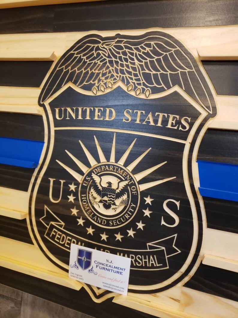 May include: Wooden wall art featuring a carved United States Federal Air Marshal badge. The badge is black with gold lettering and a blue line, set against a wood grain background. The text "United States" is visible.