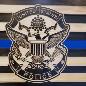 K&ouml;nnte beinhalten: Wandkunst aus Holz mit dem Emblem der United States Park Police, das einen Adler, einen Schild und ein Banner zeigt. Das Design umfasst eine blaue Linie und einen gestreiften Hintergrund aus Natur- und dunklem Holz.