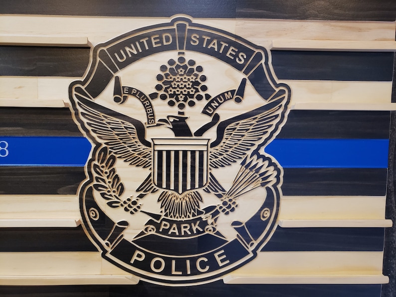 K&ouml;nnte beinhalten: Wandkunst aus Holz mit dem Emblem der United States Park Police, das einen Adler, einen Schild und ein Banner zeigt. Das Design umfasst eine blaue Linie und einen gestreiften Hintergrund aus Natur- und dunklem Holz.