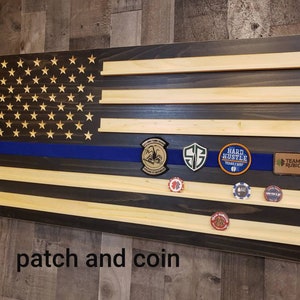 Op de afbeelding: Een Amerikaanse houten vlag met een blauwe lijn, met een patch- en muntdisplay. De vlag heeft een donkere houtkleur en lichte houten planken. De patches omvatten "Hard Hustle" en "Team Rubicon".