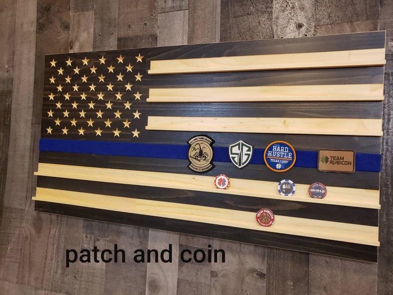 Op de afbeelding: Een Amerikaanse houten vlag met een blauwe lijn, met een patch- en muntdisplay. De vlag heeft een donkere houtkleur en lichte houten planken. De patches omvatten "Hard Hustle" en "Team Rubicon".