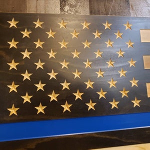 May include: Wooden American flag with carved stars and stripes. The flag features a blue stripe and a thin blue line, with the stars in a natural wood color. The background is a dark, stained wood.
