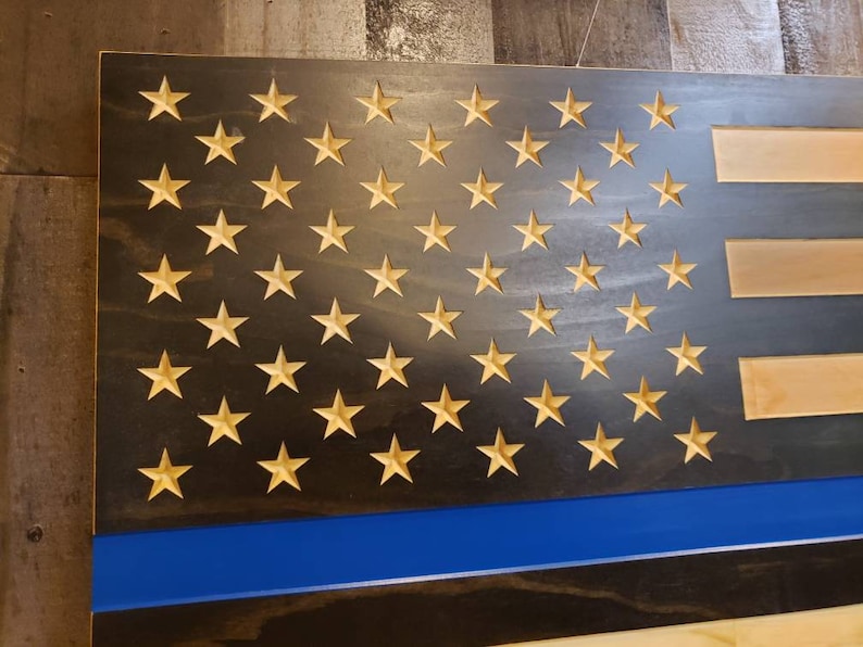 May include: Wooden American flag with carved stars and stripes. The flag features a blue stripe and a thin blue line, with the stars in a natural wood color. The background is a dark, stained wood.