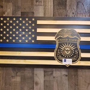 May include: Wooden American flag with a blue line and a United States Federal Marshal badge. The flag features stars and stripes, with a blue stripe representing law enforcement. The badge is intricately carved and adds a unique touch to the patriotic decor.