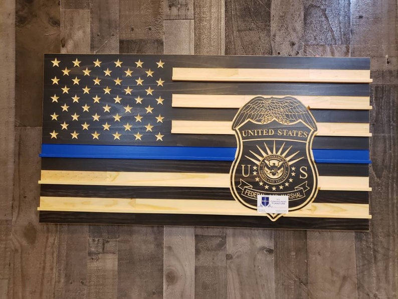 May include: Wooden American flag with a blue line and a United States Federal Marshal badge. The flag features stars and stripes, with a blue stripe representing law enforcement. The badge is intricately carved and adds a unique touch to the patriotic decor.
