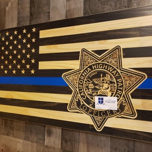 May include: A wooden American flag with a blue line, featuring a carved California Highway Patrol badge with the words "California Highway Patrol" and "Officer" in a circle. The badge is a six-pointed star with a smaller circle in the center depicting the California state seal.