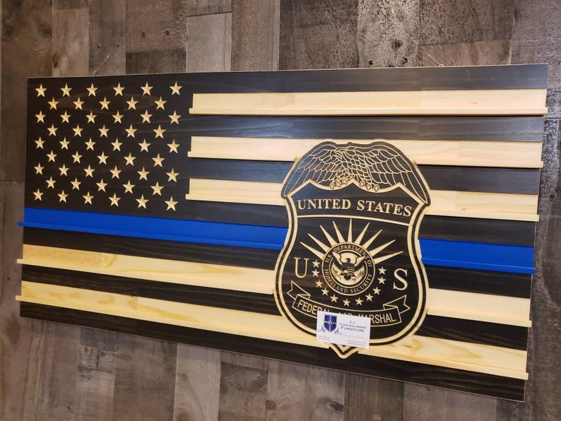 May include: Wooden American flag with a blue line and a carved United States Federal Marshal badge. The flag features stars and stripes in natural wood, black, and blue. The badge is intricately detailed with text and a seal.