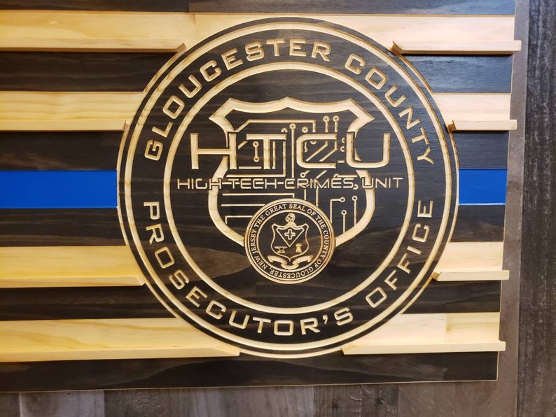 K&ouml;nnte beinhalten: Amerikanische Flagge aus Holz mit blauem Streifen und rundem Emblem. Das Emblem lautet "Gloucester County Prosecutor's Office" und "High-Tech Crimes Unit."