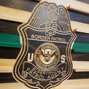 May include: Wooden CBP Border Patrol badge with a detailed eagle design. The badge includes the text "U.S. Department of Homeland Security" and "Supervisory Patrol Agent". The badge is set against a wooden background with green and black stripes.