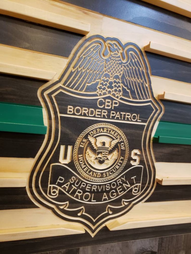 May include: Wooden CBP Border Patrol badge with a detailed eagle design. The badge includes the text "U.S. Department of Homeland Security" and "Supervisory Patrol Agent". The badge is set against a wooden background with green and black stripes.