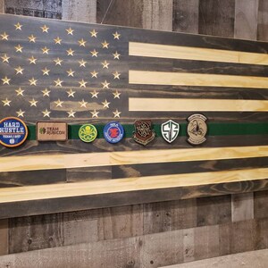 Op de afbeelding: Een Amerikaanse vlag van hout met een groene streep in het midden. De vlag heeft een verweerde uitstraling en is versierd met verschillende patches, waaronder "Hard Hustle", "Team Rubicon" en een schedel met gekruiste beenderen.