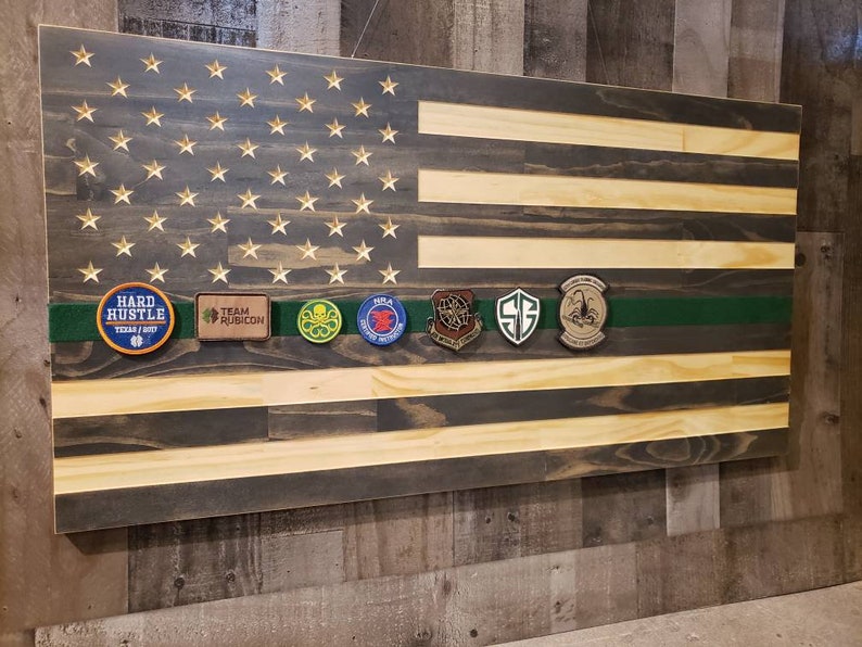 Op de afbeelding: Een Amerikaanse vlag van hout met een groene streep in het midden. De vlag heeft een verweerde uitstraling en is versierd met verschillende patches, waaronder "Hard Hustle", "Team Rubicon" en een schedel met gekruiste beenderen.