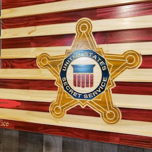 May include: A wooden American flag with the United States Secret Service emblem carved into the center. The flag is stained in red, white, and blue.