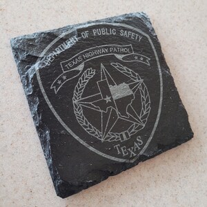 May include: Black slate coaster with a white engraved design of the Texas Department of Public Safety logo. The logo features a five-pointed star with a smaller star inside, surrounded by a wreath and the words "Texas Highway Patrol".