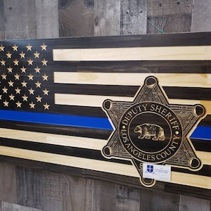 May include: A wooden American flag with a blue line and a Los Angeles County Sheriff's star badge in the center. The flag is made of dark and light wood.