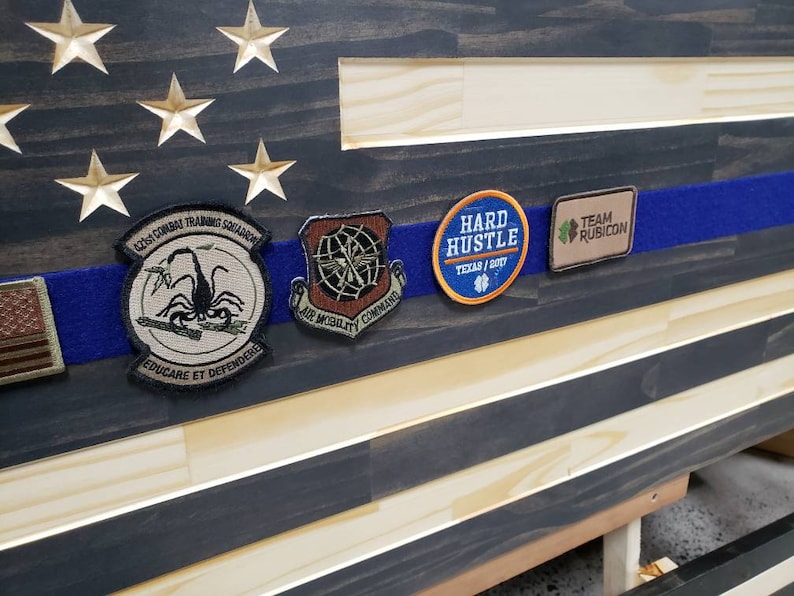 Op de afbeelding: Een Amerikaanse vlag van hout met een blauwe streep in het midden. Op de blauwe streep zitten verschillende patches, waaronder een patch met een schorpioen, een patch met een spinnenweb, een patch met de tekst "Hard Hustle" en een patch met de tekst "Team Rubicon".