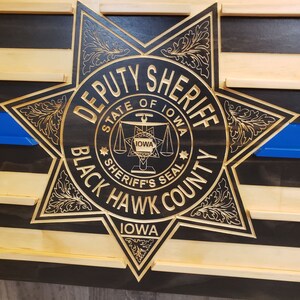 K&ouml;nnte beinhalten: Holzwandkunst mit einem geschnitzten Deputy Sheriff-Abzeichen aus Black Hawk County, Iowa. Das Abzeichen ist goldfarben und befindet sich vor einem schwarz-naturfarbenen Holzstreifenhintergrund mit einer blauen Linie.