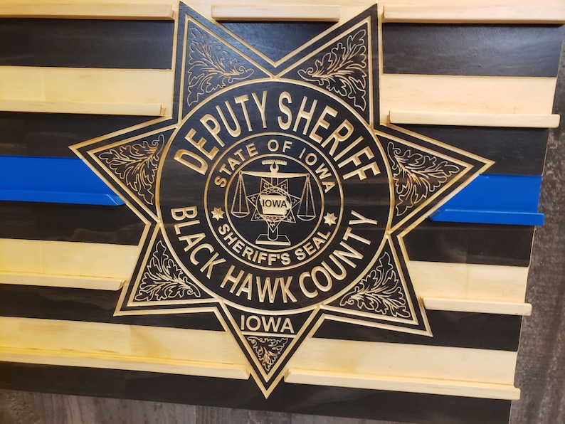 K&ouml;nnte beinhalten: Holzwandkunst mit einem geschnitzten Deputy Sheriff-Abzeichen aus Black Hawk County, Iowa. Das Abzeichen ist goldfarben und befindet sich vor einem schwarz-naturfarbenen Holzstreifenhintergrund mit einer blauen Linie.
