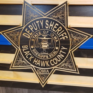 K&ouml;nnte beinhalten: Holzwandkunst mit einem geschnitzten Deputy Sheriff-Abzeichen aus Black Hawk County, Iowa. Das Abzeichen ist goldfarben und befindet sich vor einem schwarz-naturfarbenen Holzstreifenhintergrund mit einer blauen Linie.