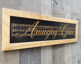 Puede incluir: Un letrero enmarcado de madera rústica con fondo negro y letras doradas que dice "Amazing Grace". El letrero presenta notas musicales impresas en dorado sobre el fondo negro.