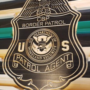 May include: Wooden plaque featuring a CBP Border Patrol badge design. The badge is black with a detailed border and engraved text, including "CBP Border Patrol," "U.S. Department of Homeland Security," and "Patrol Agent." An eagle tops the badge.